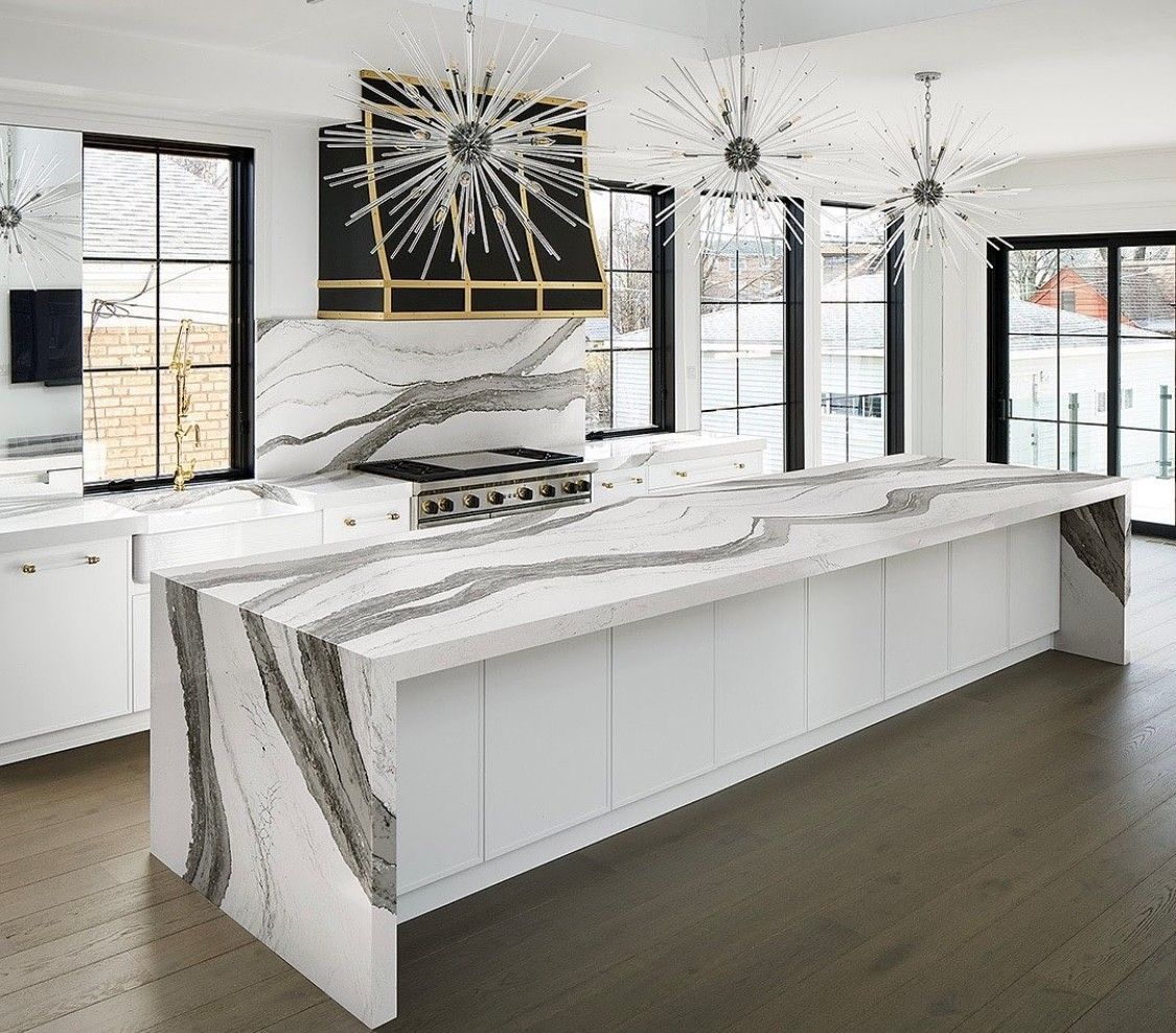 Modern white kitchen with large island and marble countertops, black windows, and decorative light fixtures.