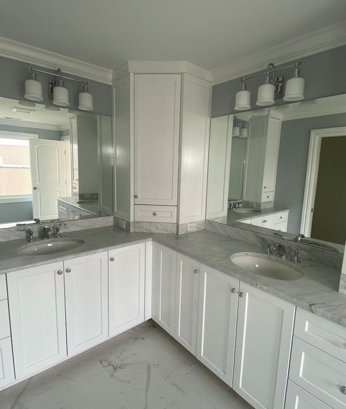 White bathroom vanity with two sinks, mirrors, and overhead lighting. Corner cabinet, light blue walls, and marble countertops.