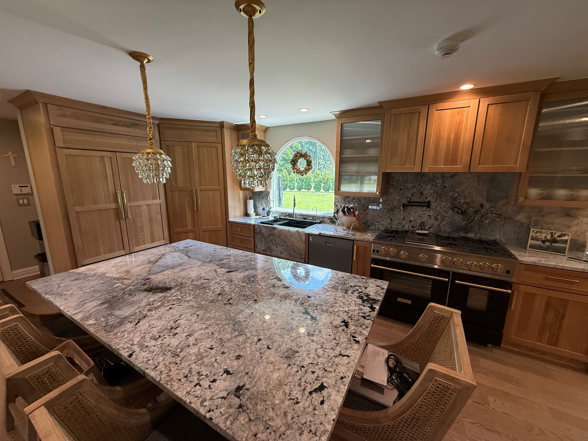 Spacious kitchen with a large granite island, wood cabinets, and a stove. Two chandeliers hang overhead.