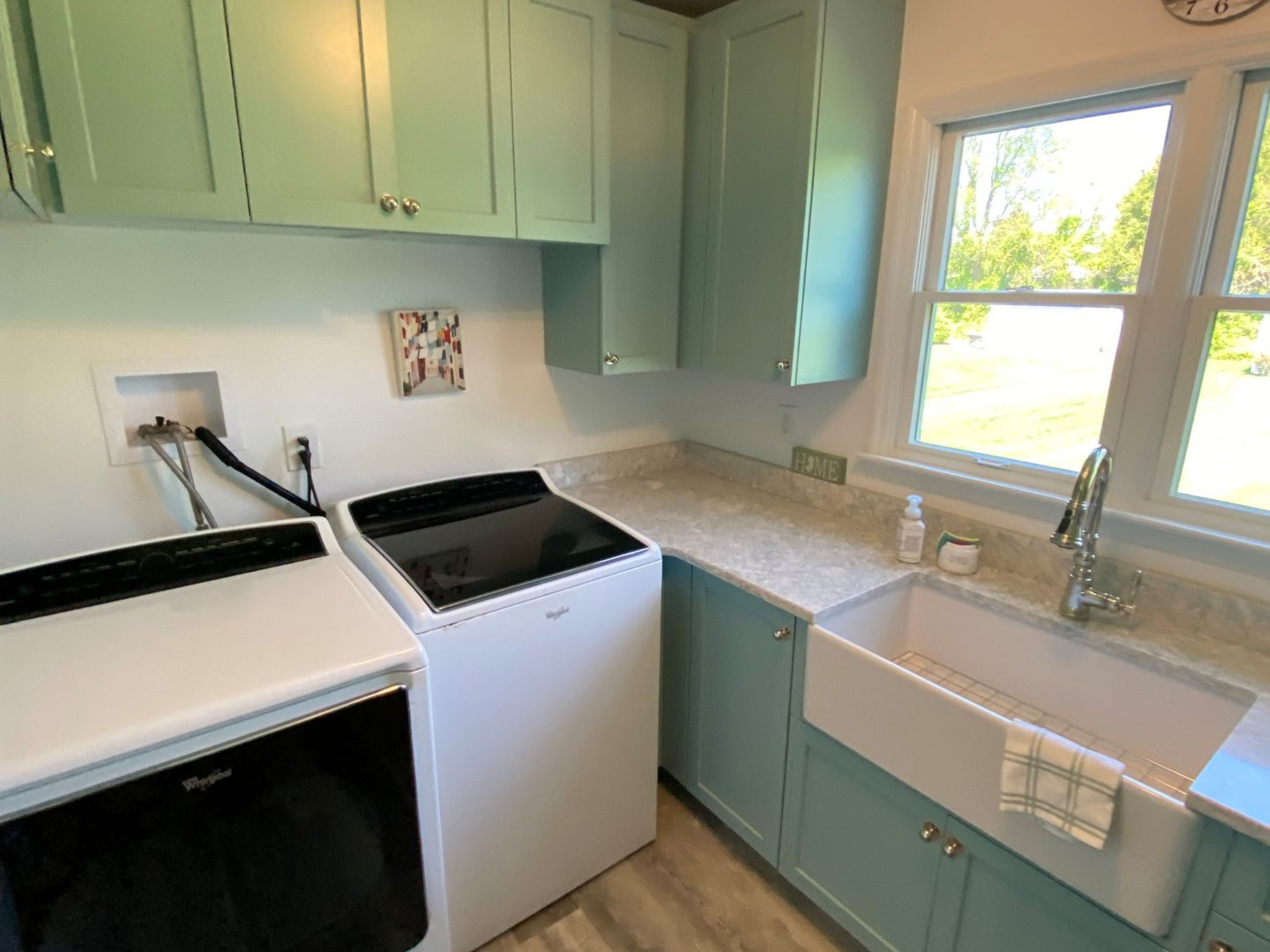 Laundry room with light blue cabinets, white appliances, and a white farmhouse sink by a window.