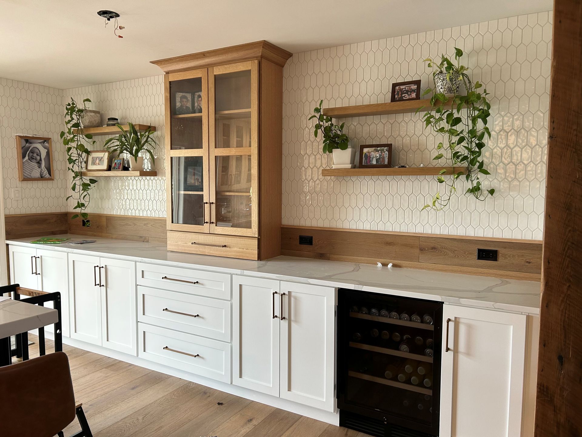 A kitchen with white cabinets, wood accents, and floating shelves. Wine fridge and a glass-front cabinet.