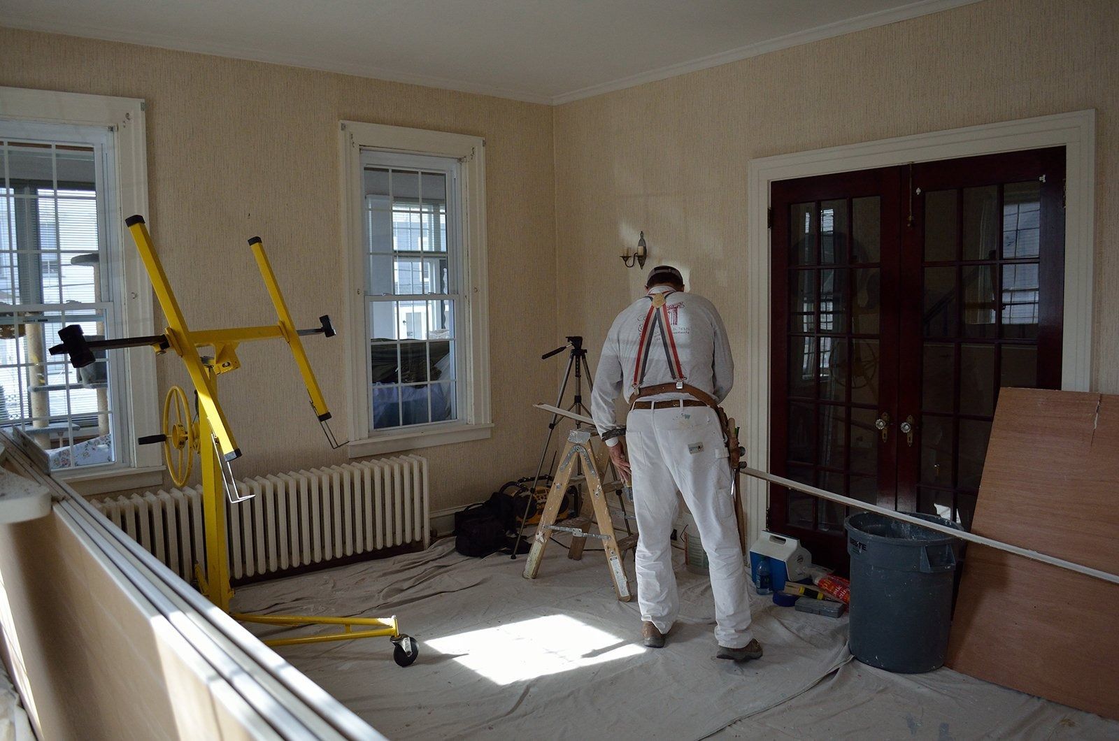 Man in white overalls working on wall; room with windows, drywall lift.