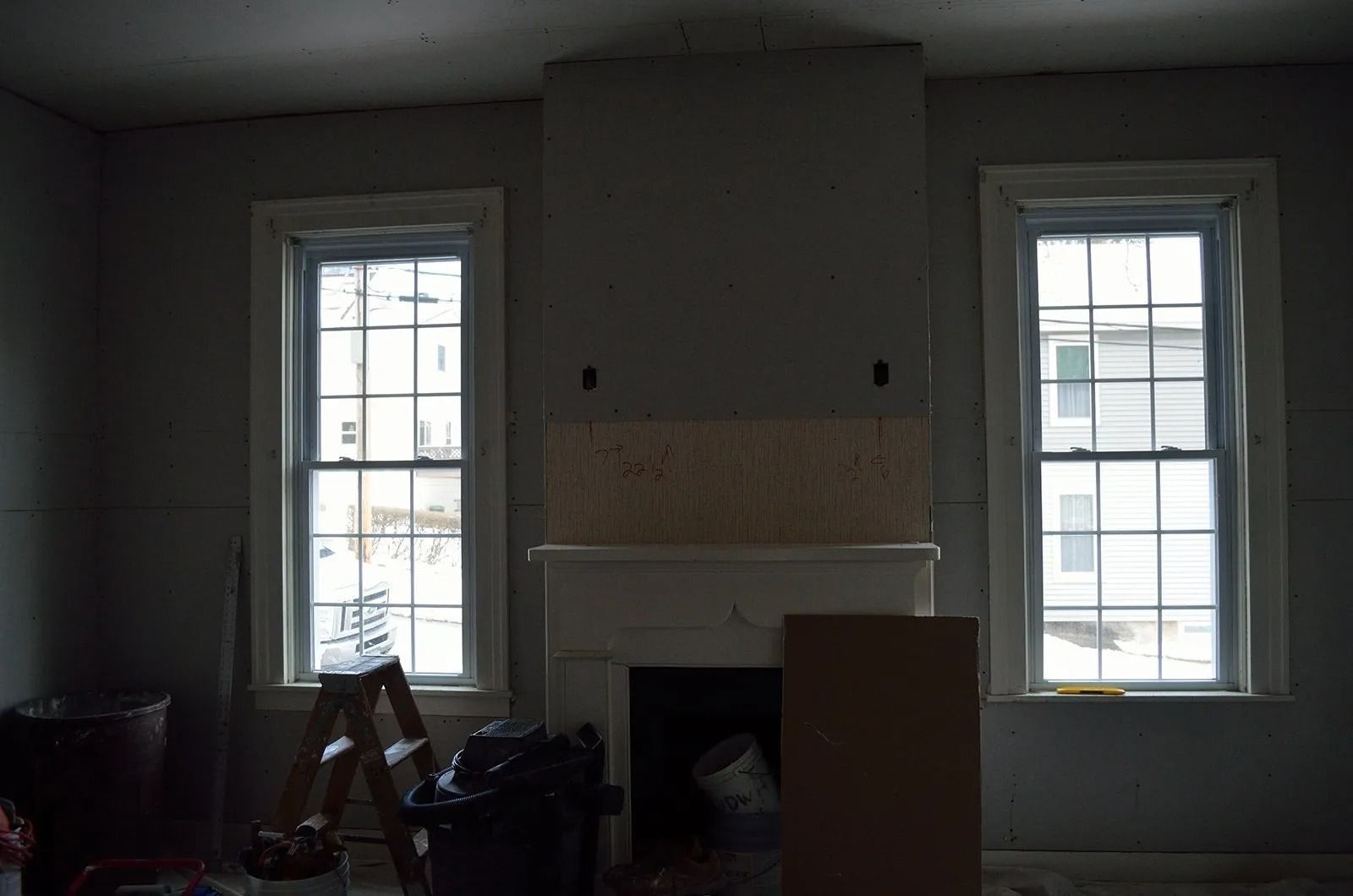 Interior view of a room under renovation, with two windows flanking a fireplace. Gray walls and natural light.