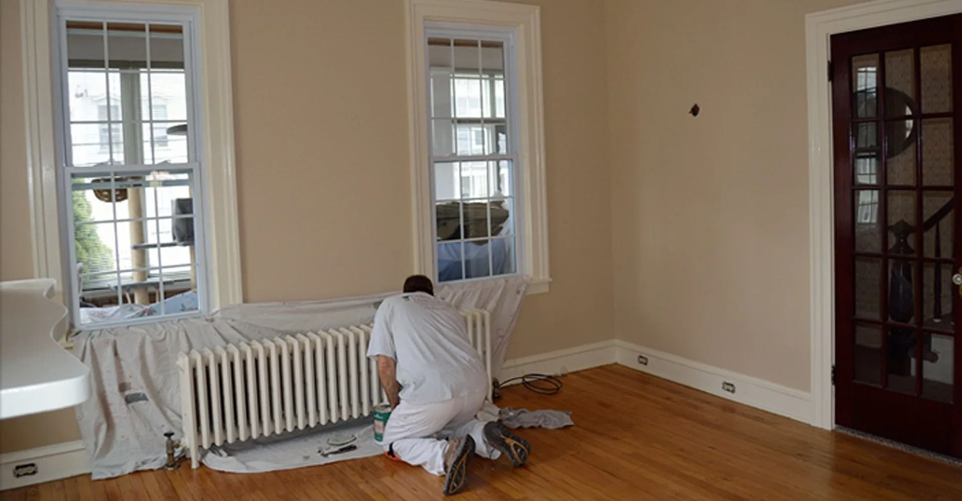 Person in white overalls kneeling, painting wall in a room with windows and wood floor.