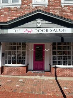 Storefront of The Pink Door Salon with a bright pink door and white awning on a brick street