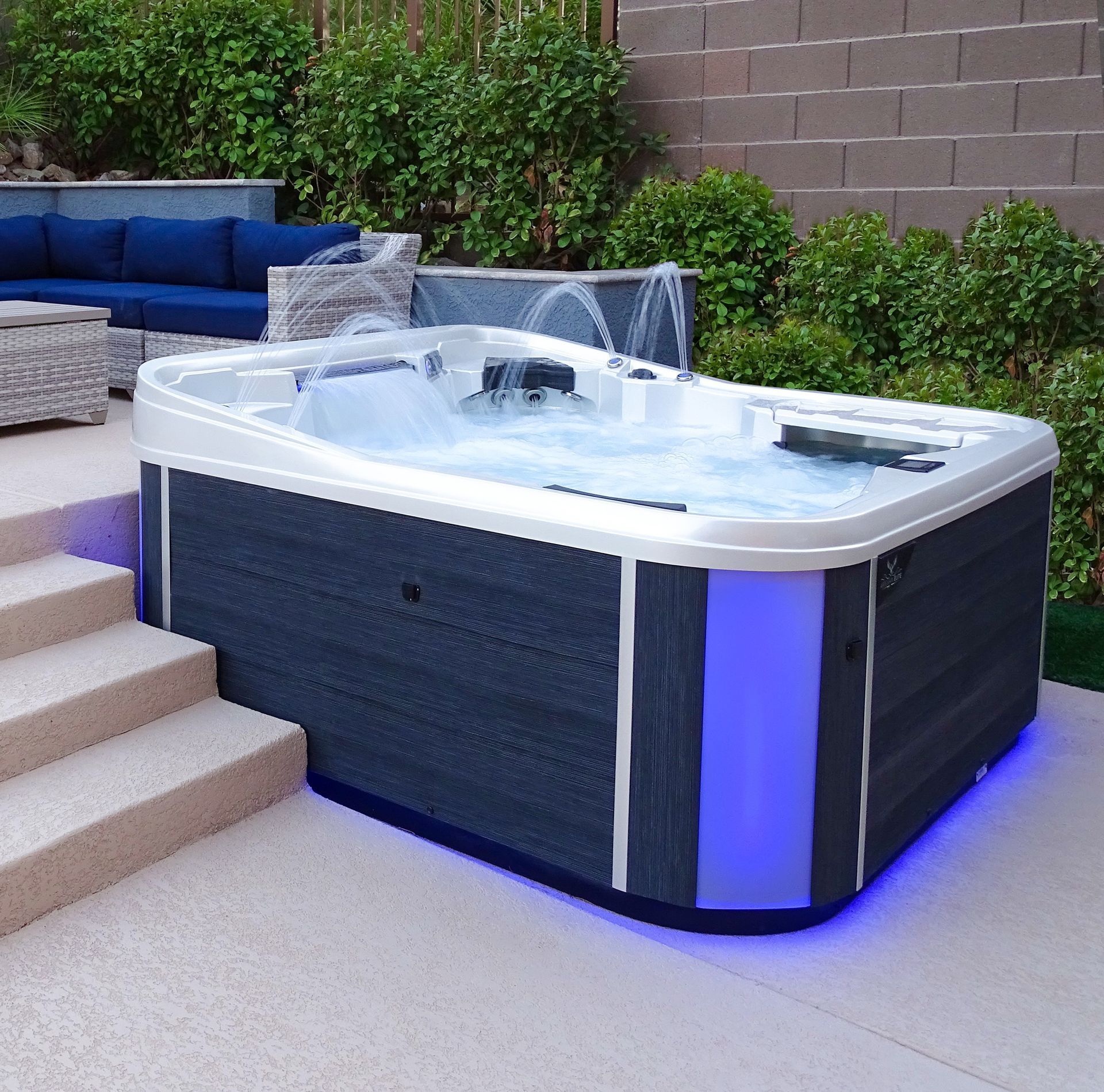 Outdoor hot tub with blue LED lighting, steps, and patio seating beside greenery and a brick wall