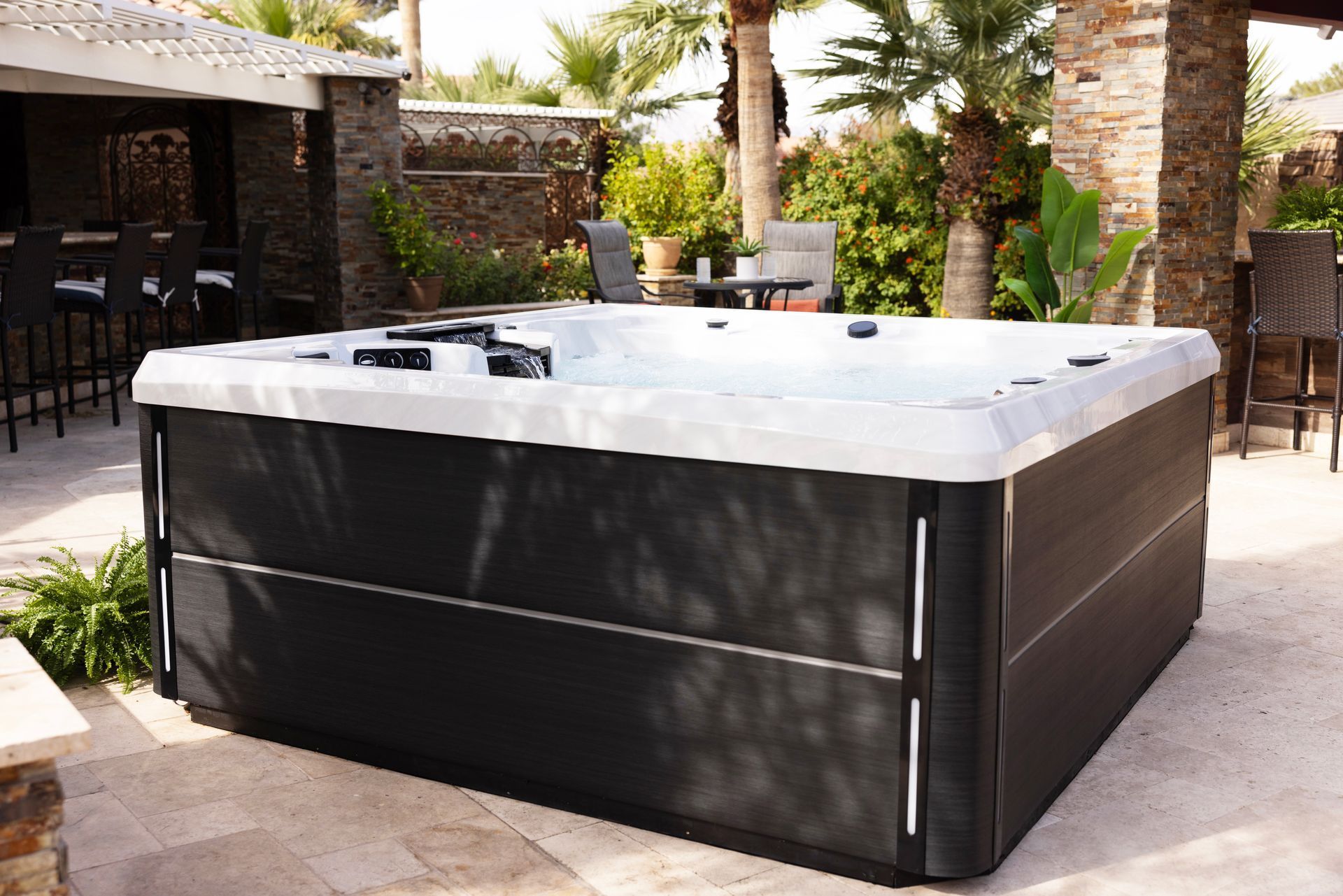Large black outdoor hot tub on a patio in a sunny backyard with stone walls and palm trees
