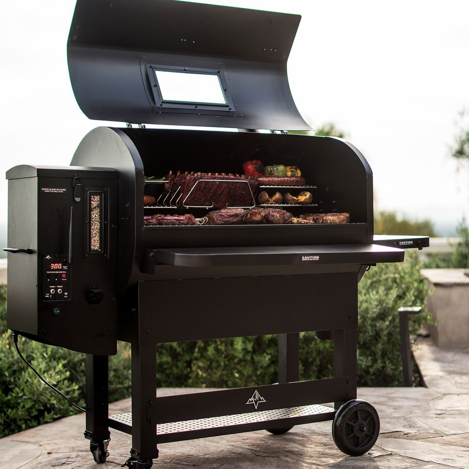 Black pellet grill with lid open, cooking meat on grates outdoors on a patio