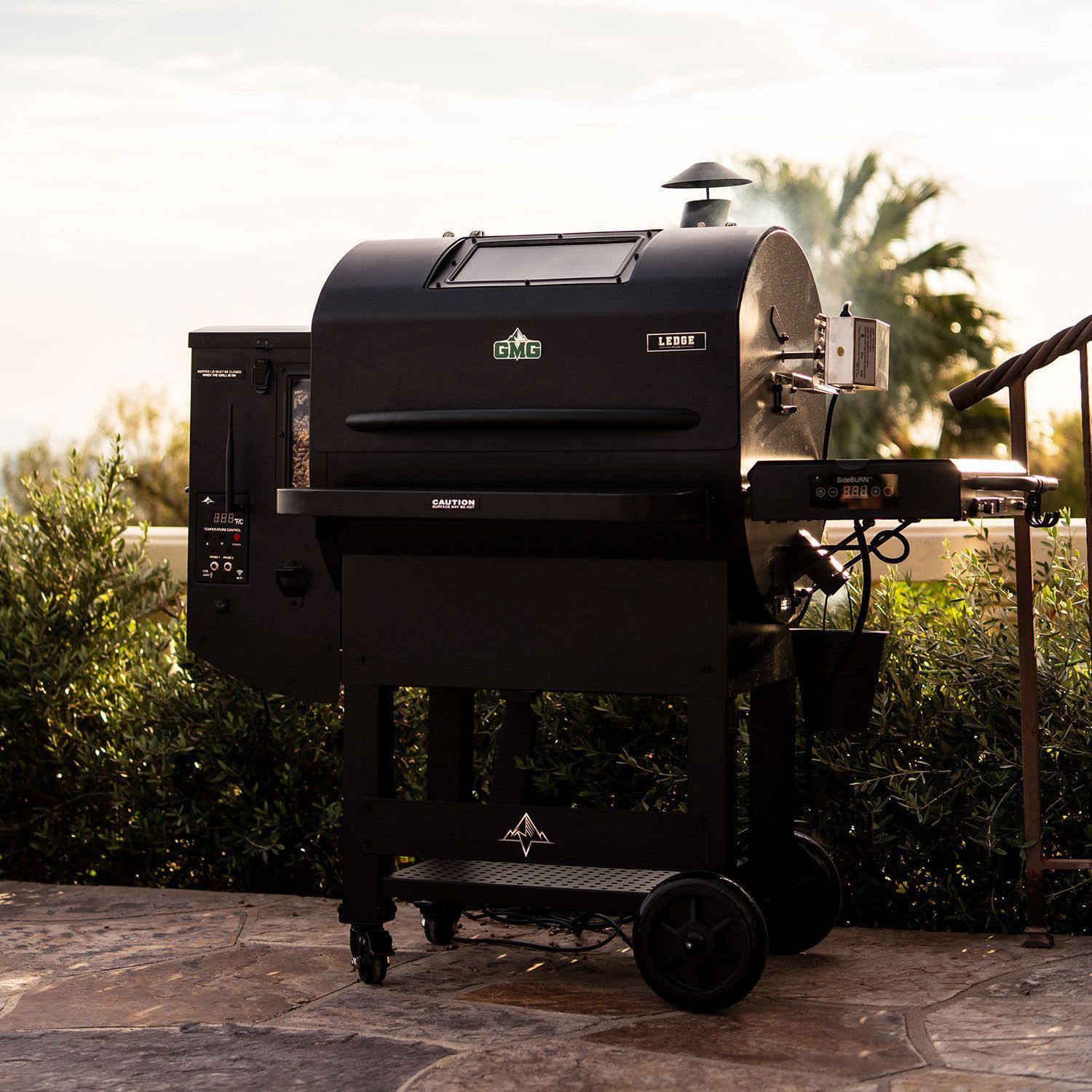 Black pellet grill on a patio with greenery and palm trees in the background