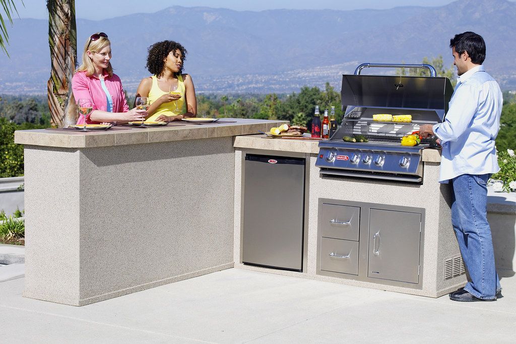 Three people chat beside an outdoor patio grill with mountains in the background.