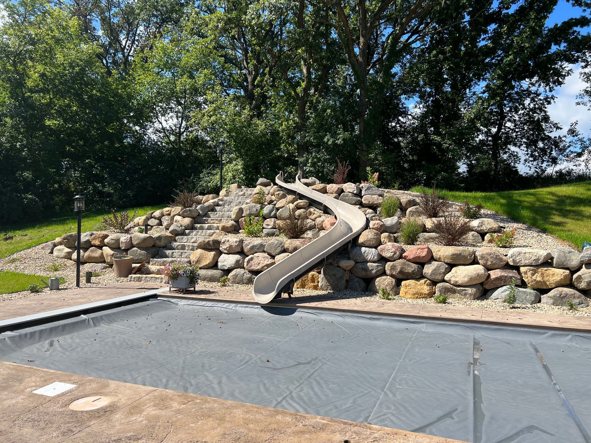 Stone drainage channel on a landscaped hill above a covered pool, with trees in the background