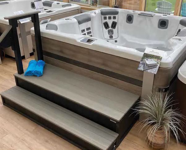 Hot tub with wooden steps and gray siding in a showroom, with blue towels and a potted plant nearby