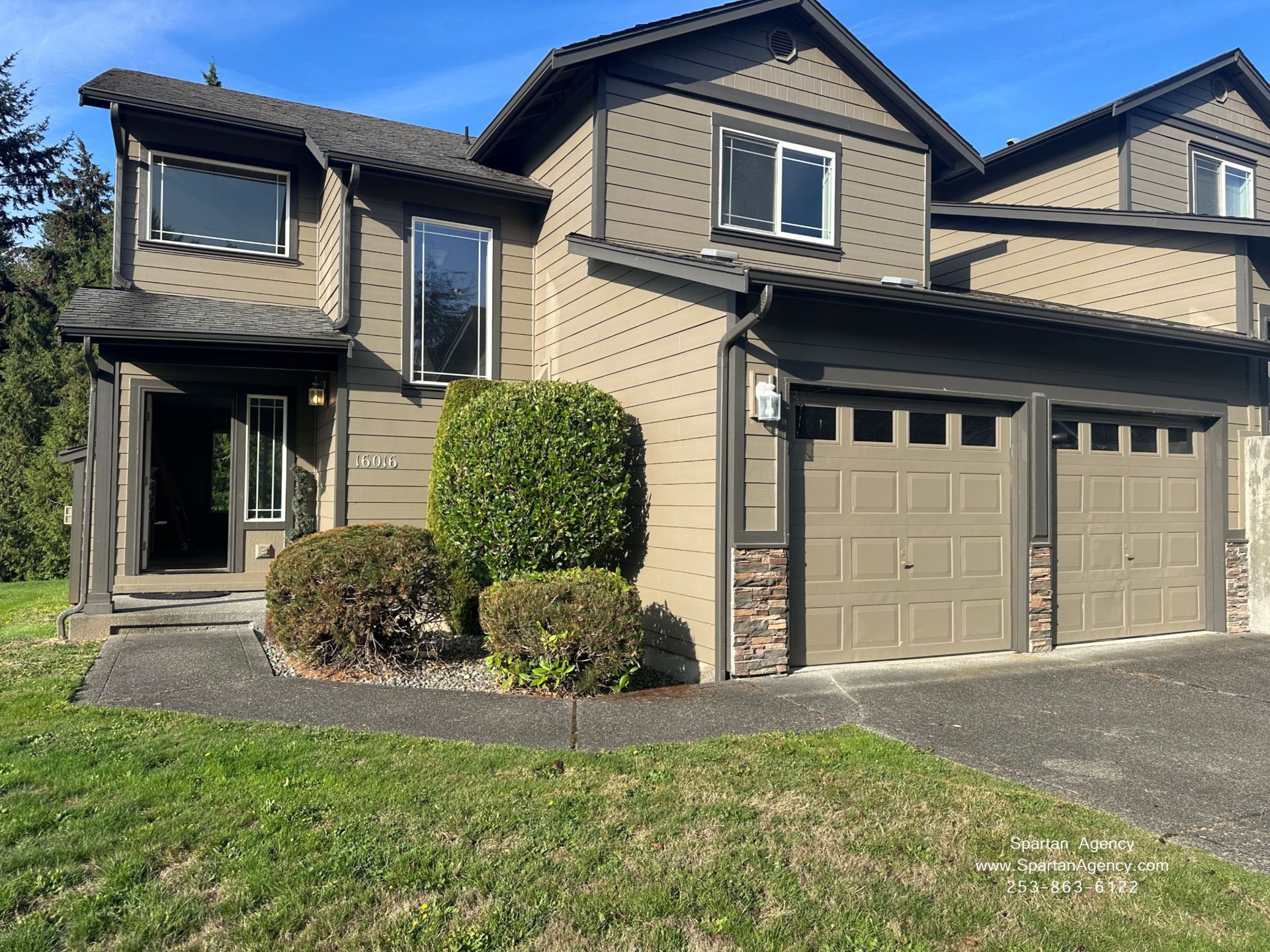 Suburban two-story house with tan siding, gray garage, and trimmed shrubs in front yard