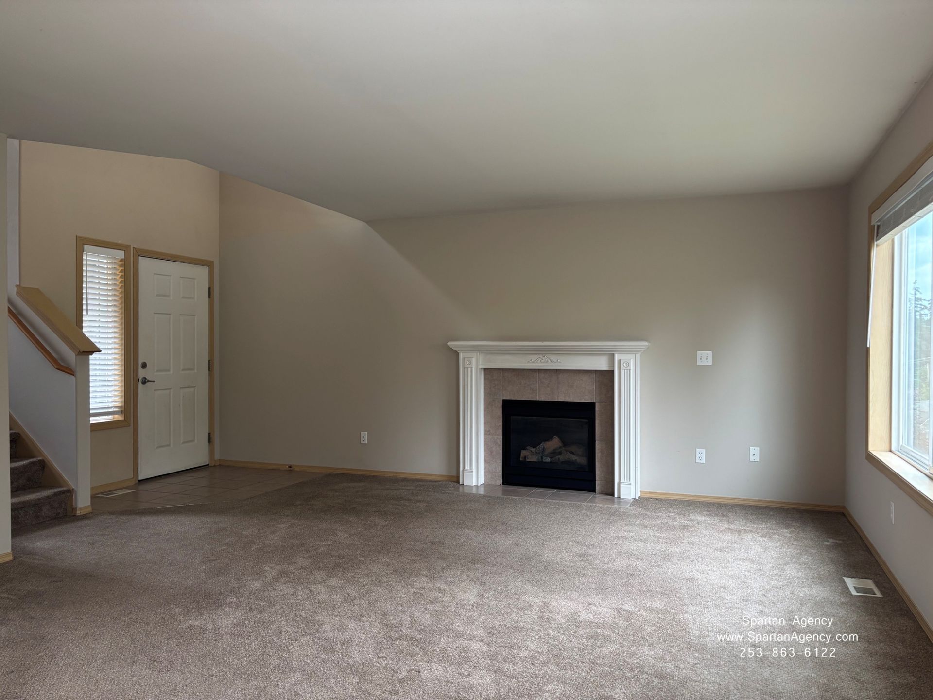 Empty living room with carpet, fireplace, stairs, and large windows in neutral tones