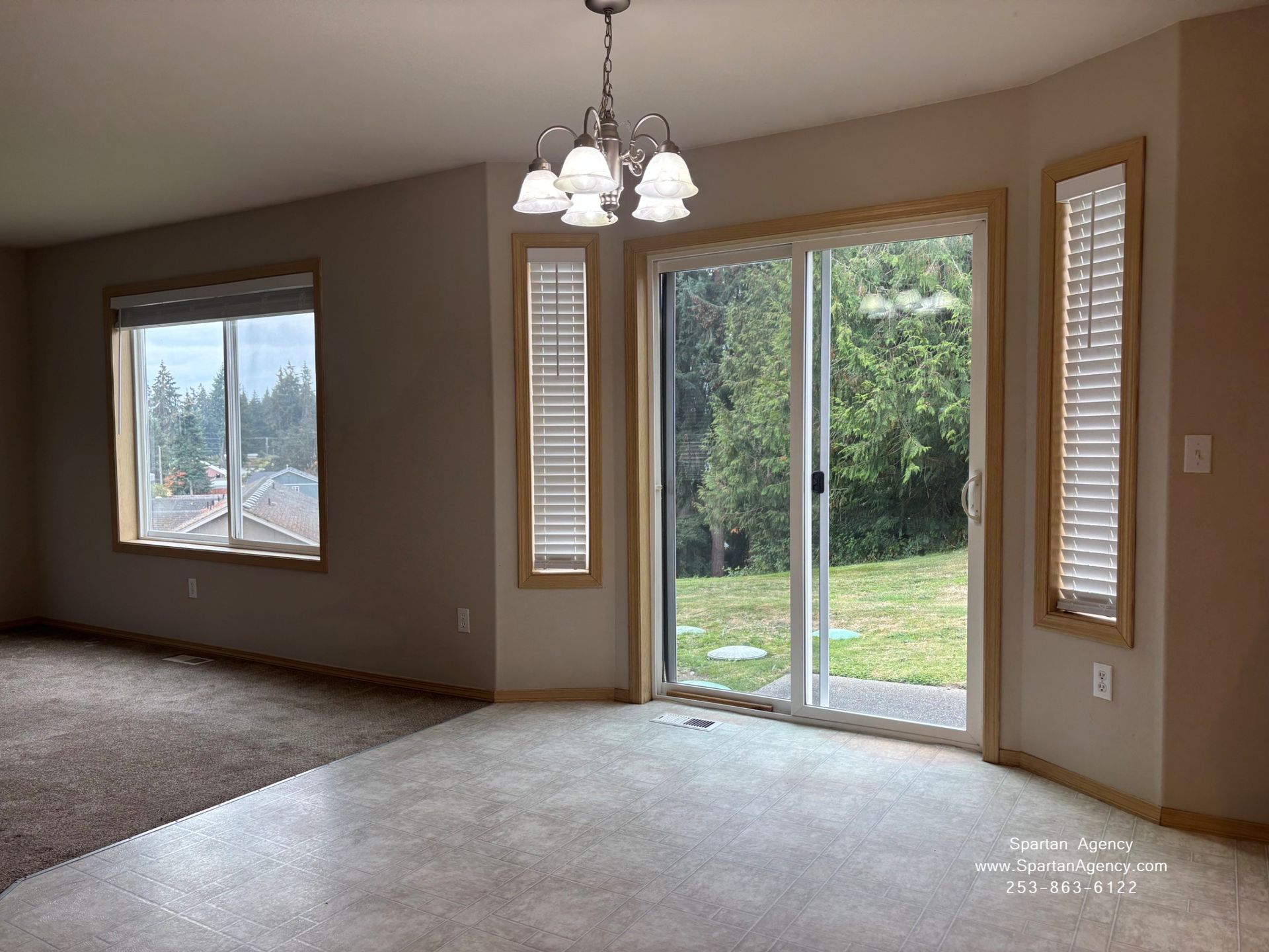Empty living-dining room with carpet, chandelier, large window, and sliding glass door to a backyard.