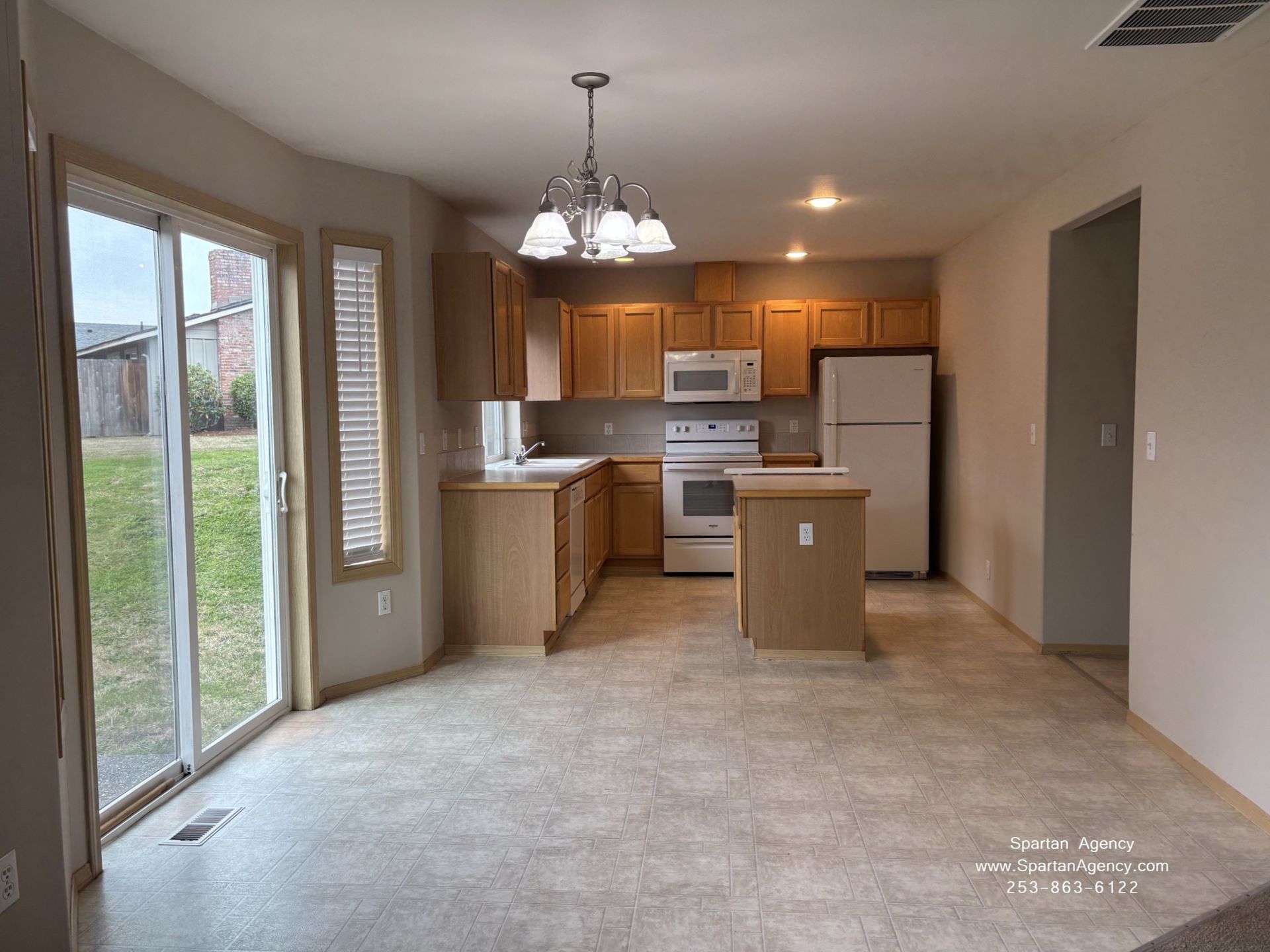 Empty beige living room opening to a small kitchen with wood cabinets, white appliances, and a glass door to the yard