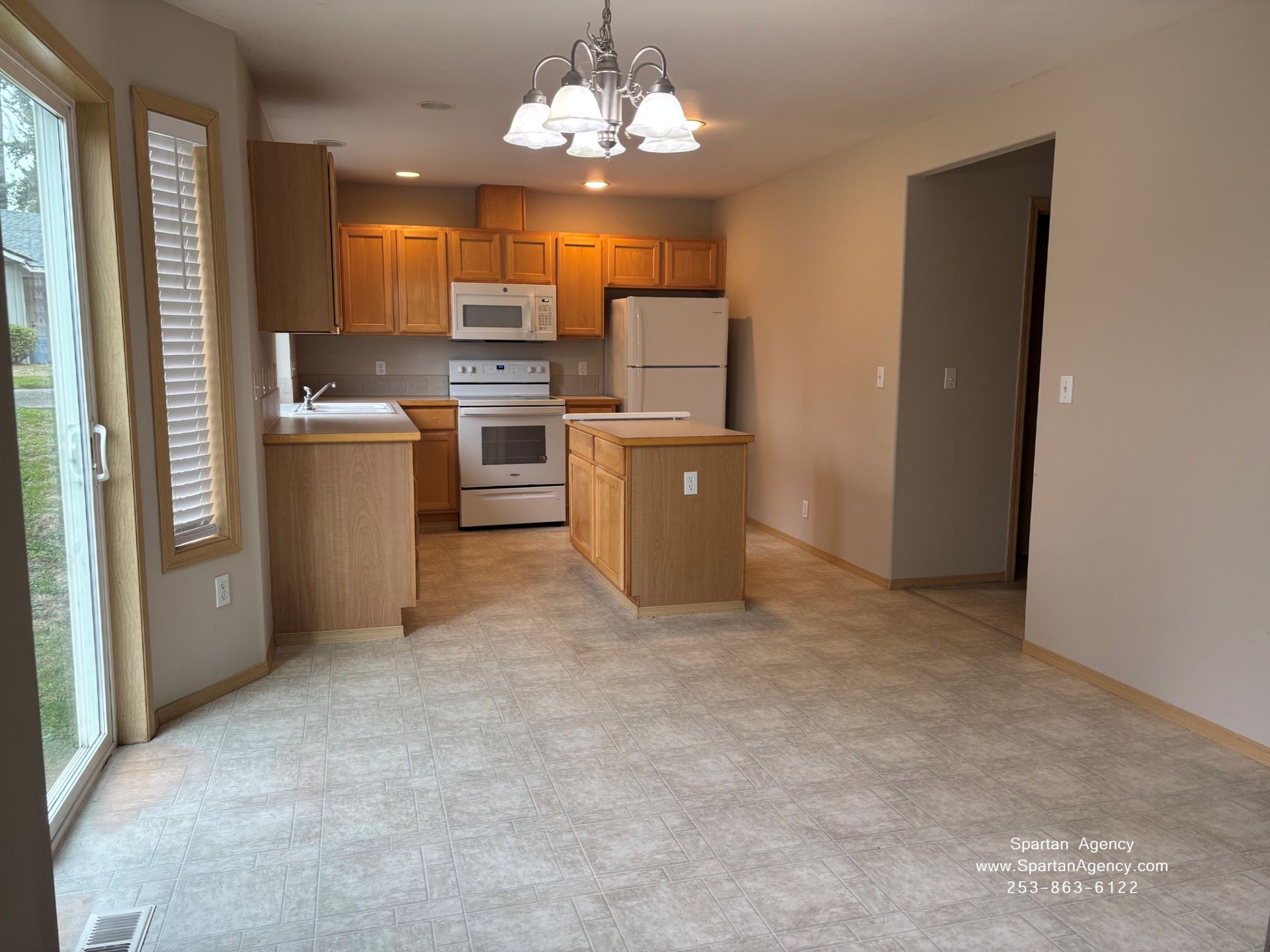 Empty apartment living area with kitchen, white appliances, wood cabinets, and a hanging light fixture