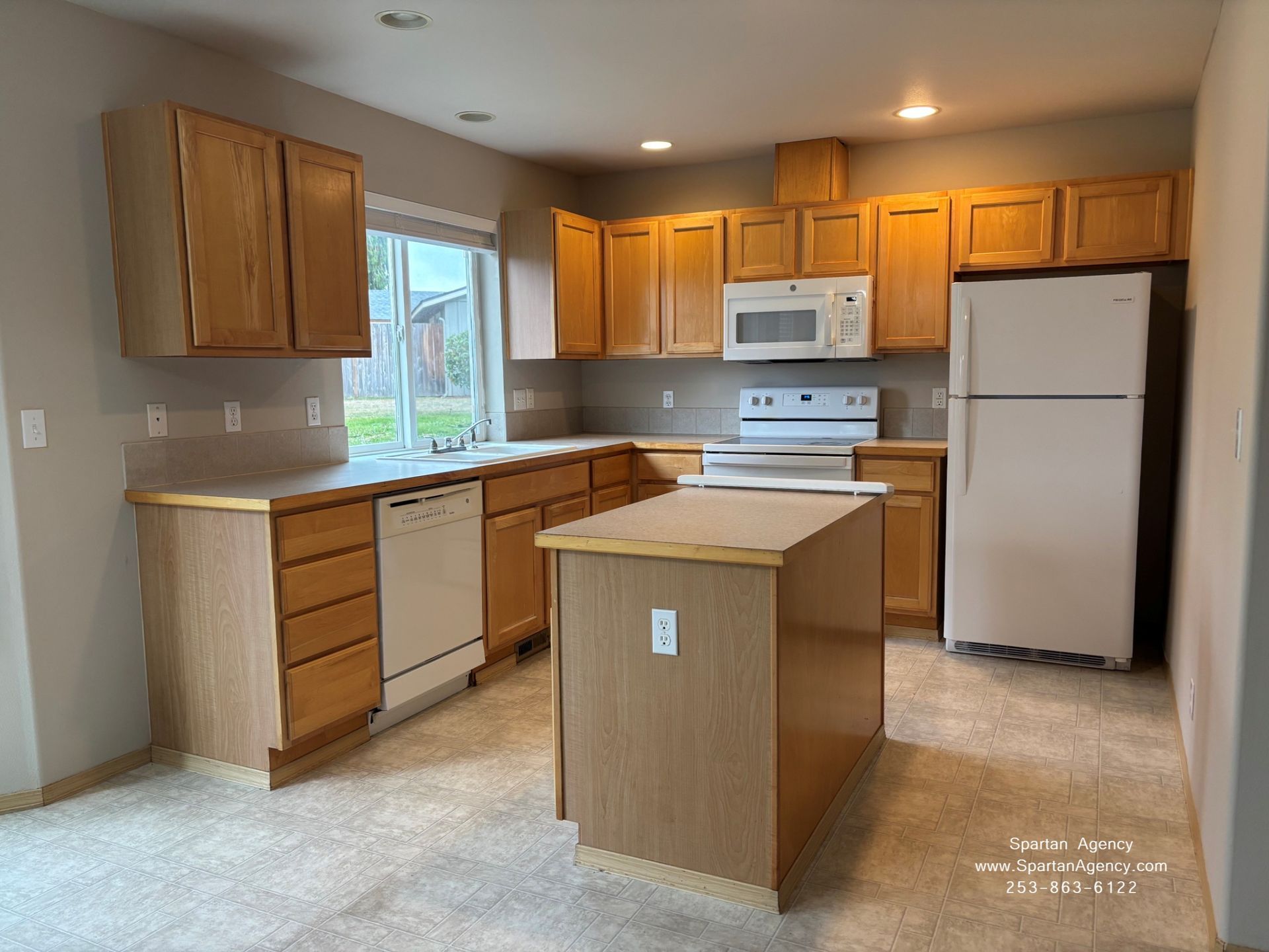 Empty kitchen with light wood cabinets, white appliances, island, and beige carpet.