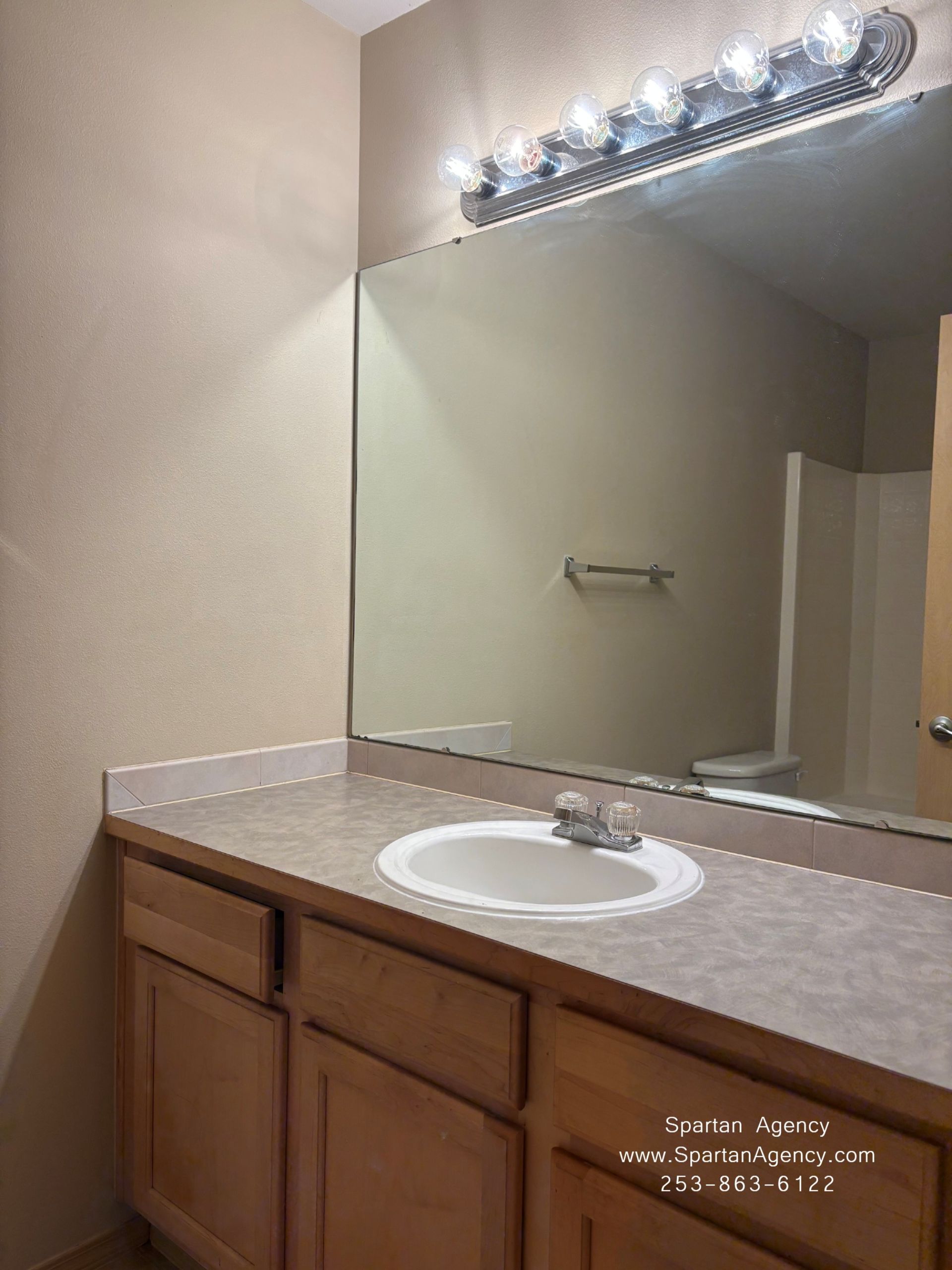 Bathroom vanity with beige countertop, wood cabinets, and large mirror under bright vanity lights.
