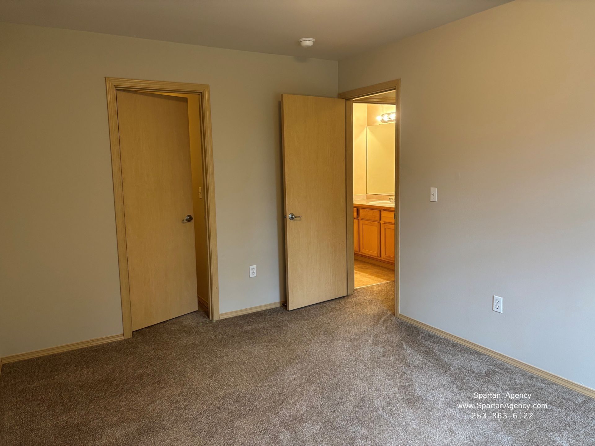 Empty beige bedroom with carpet, two wooden doors, and a doorway to a lit bathroom