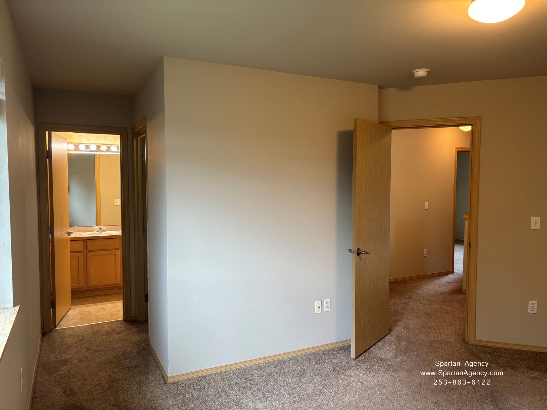 Empty bedroom with beige walls, carpeted floor, open doors, and a bathroom vanity visible in the hallway.