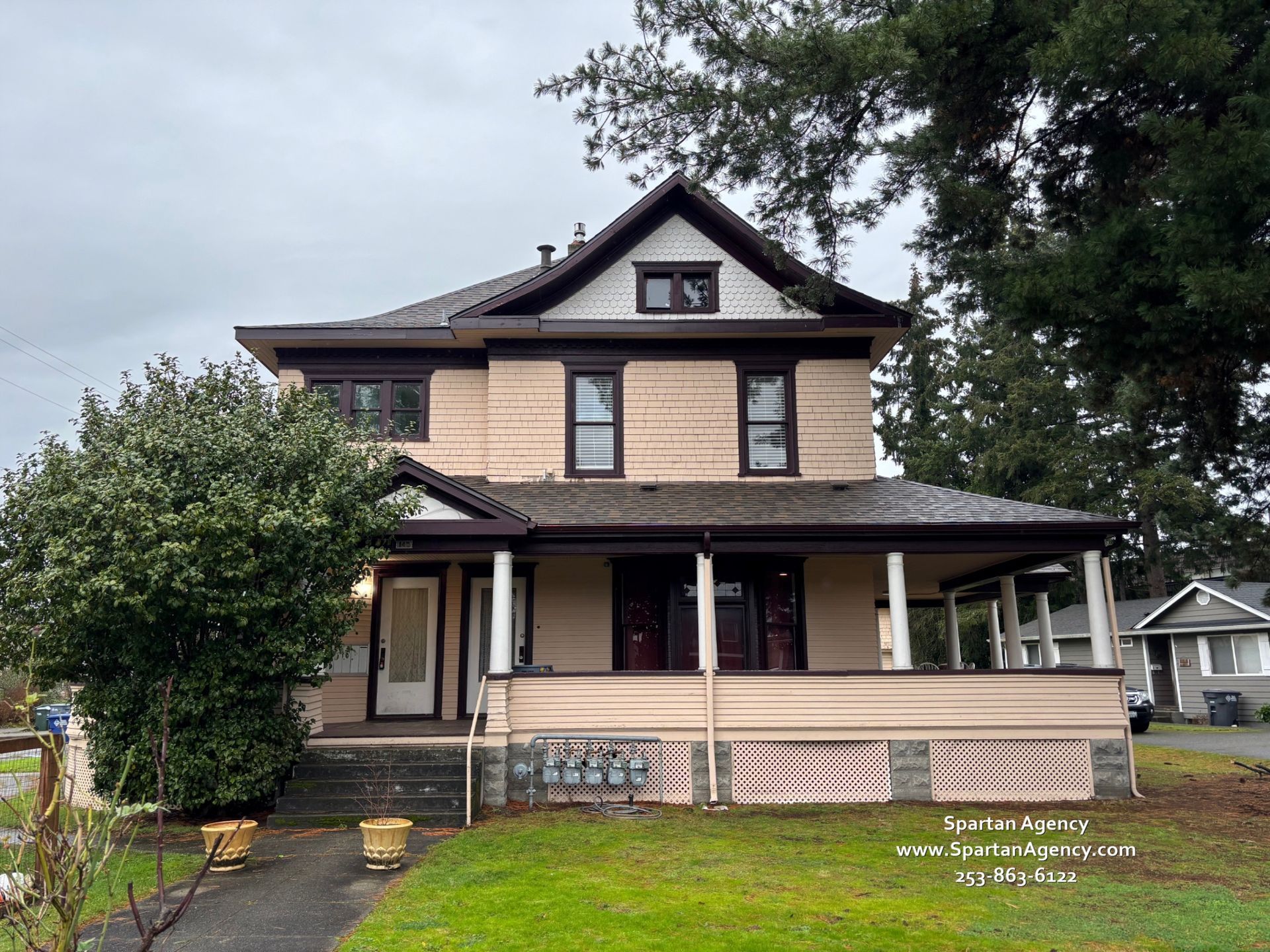 Two-story beige house with a dark roof and front porch, surrounded by trees and a lawn.