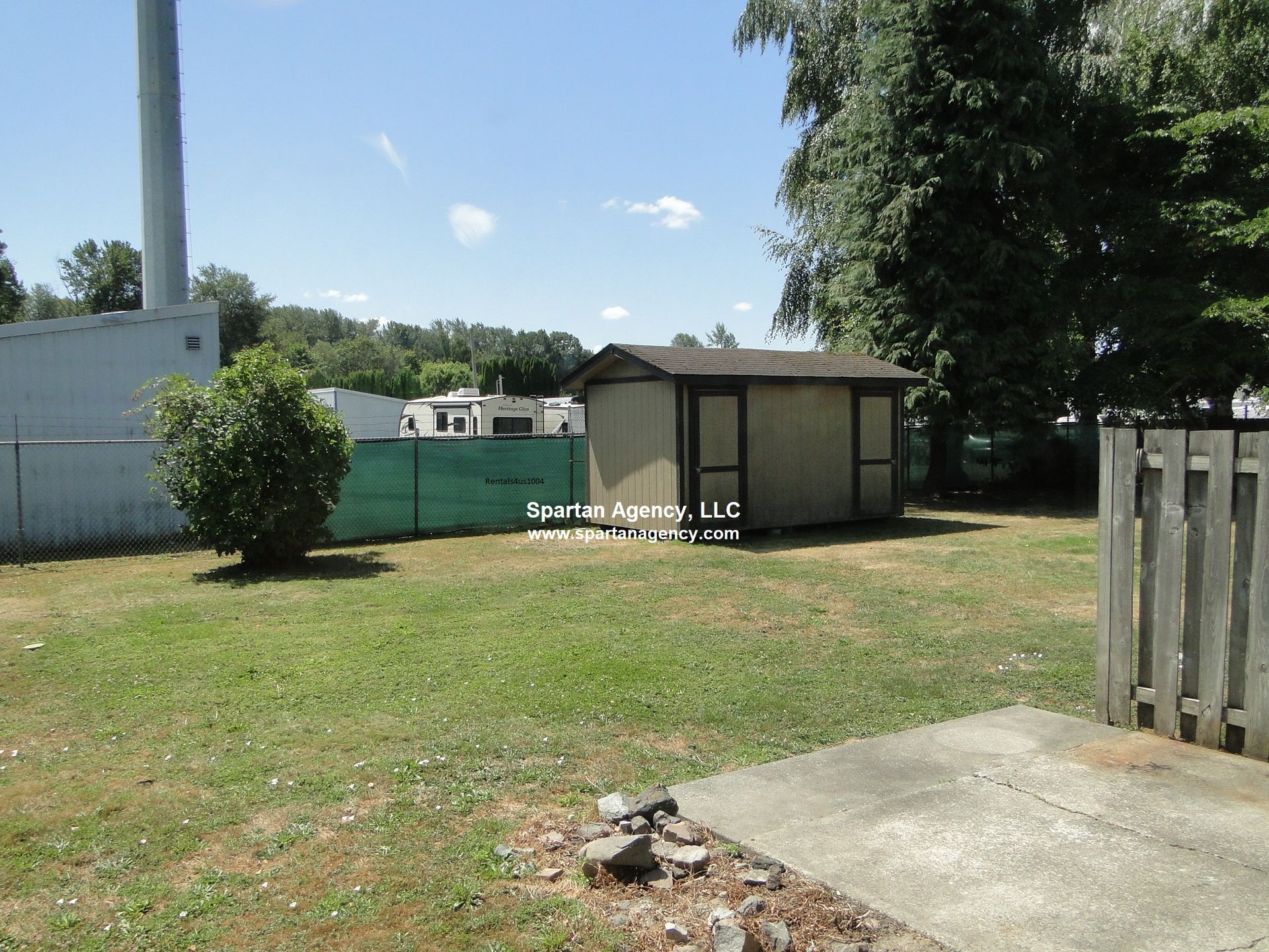 A backyard with a shed, green fence, and grass. Bright sky with trees in the background.