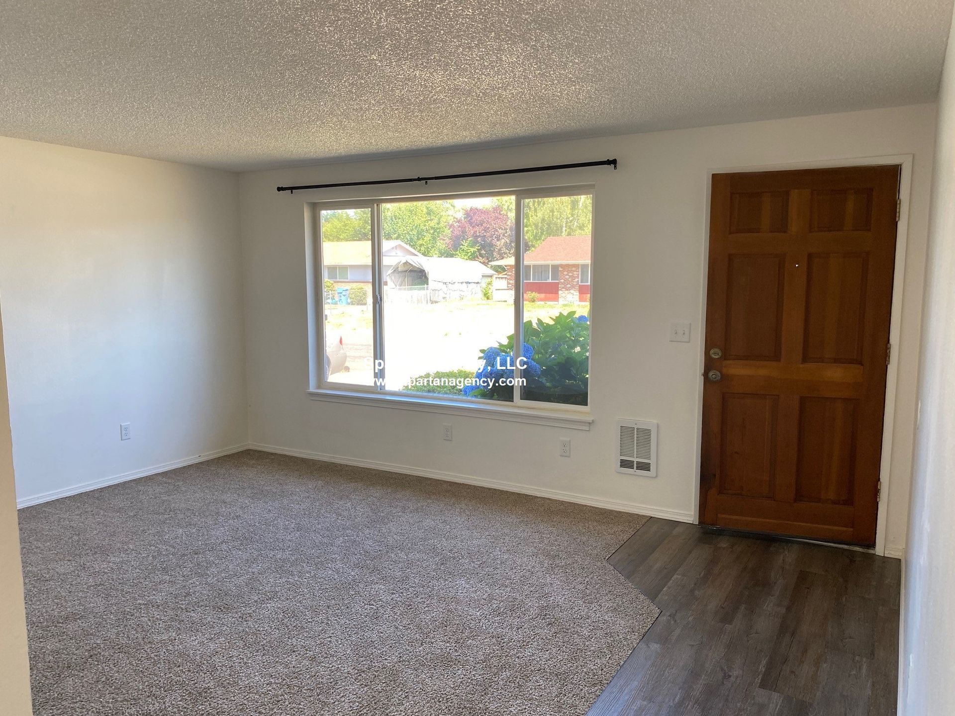 Empty living room: beige carpet, large window, brown front door, white walls, and popcorn ceiling.