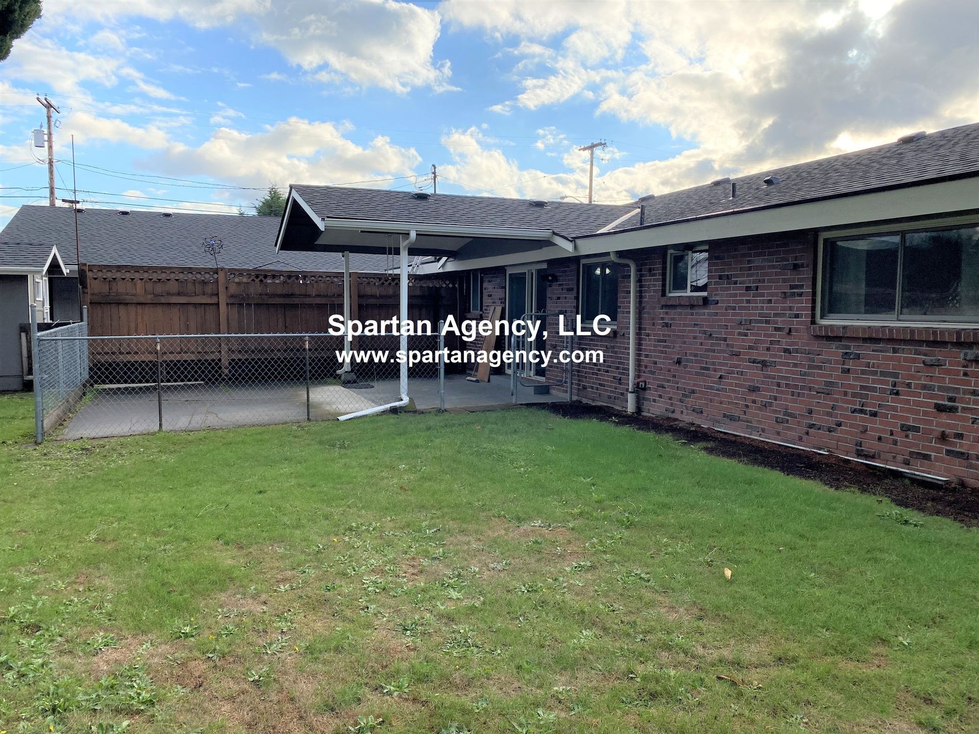 Backyard with a brick house, concrete patio, and a grassy lawn under a cloudy sky.