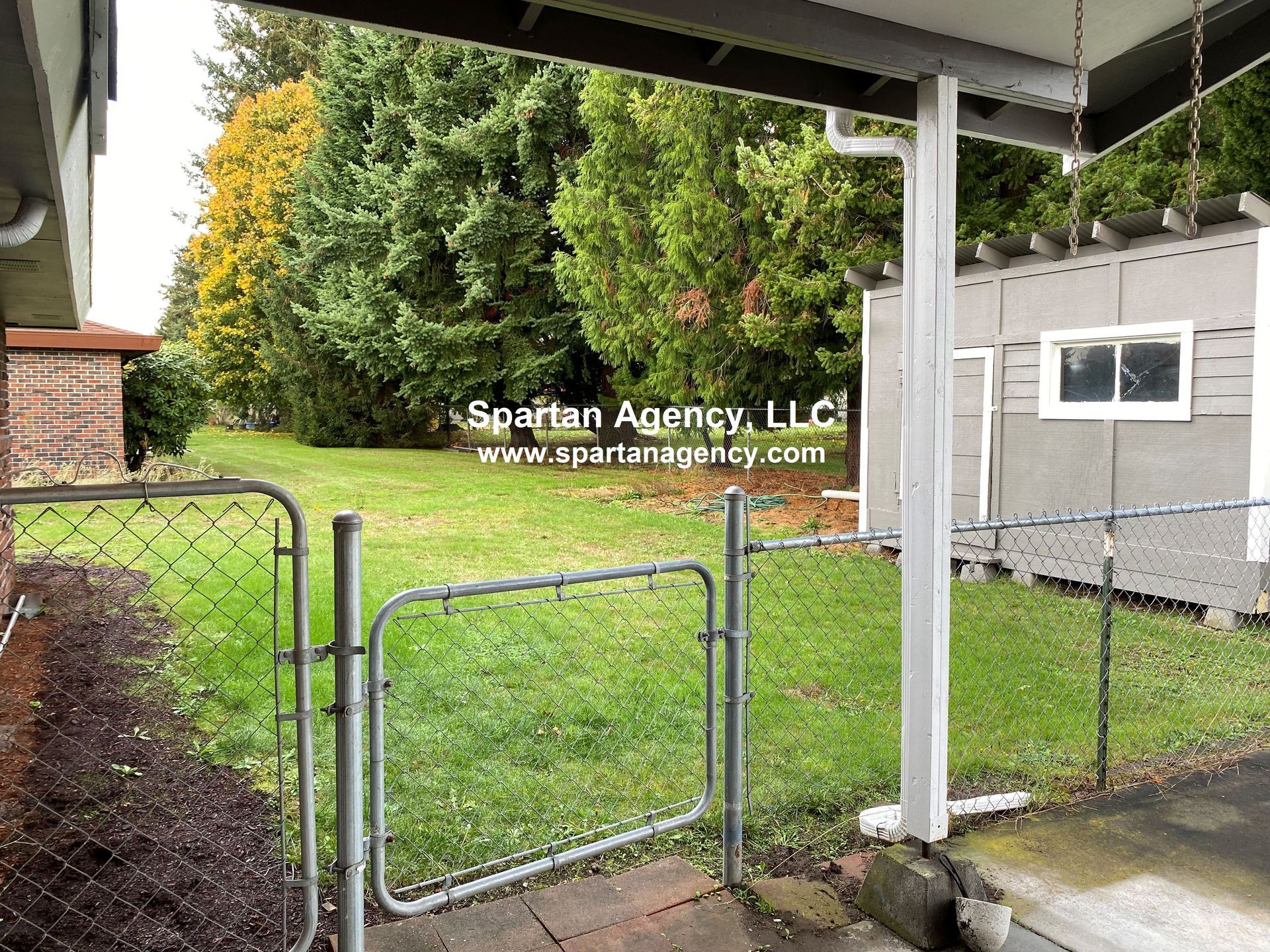 A fenced backyard with a gate, shed, and lush green grass. The view is from a covered porch.
