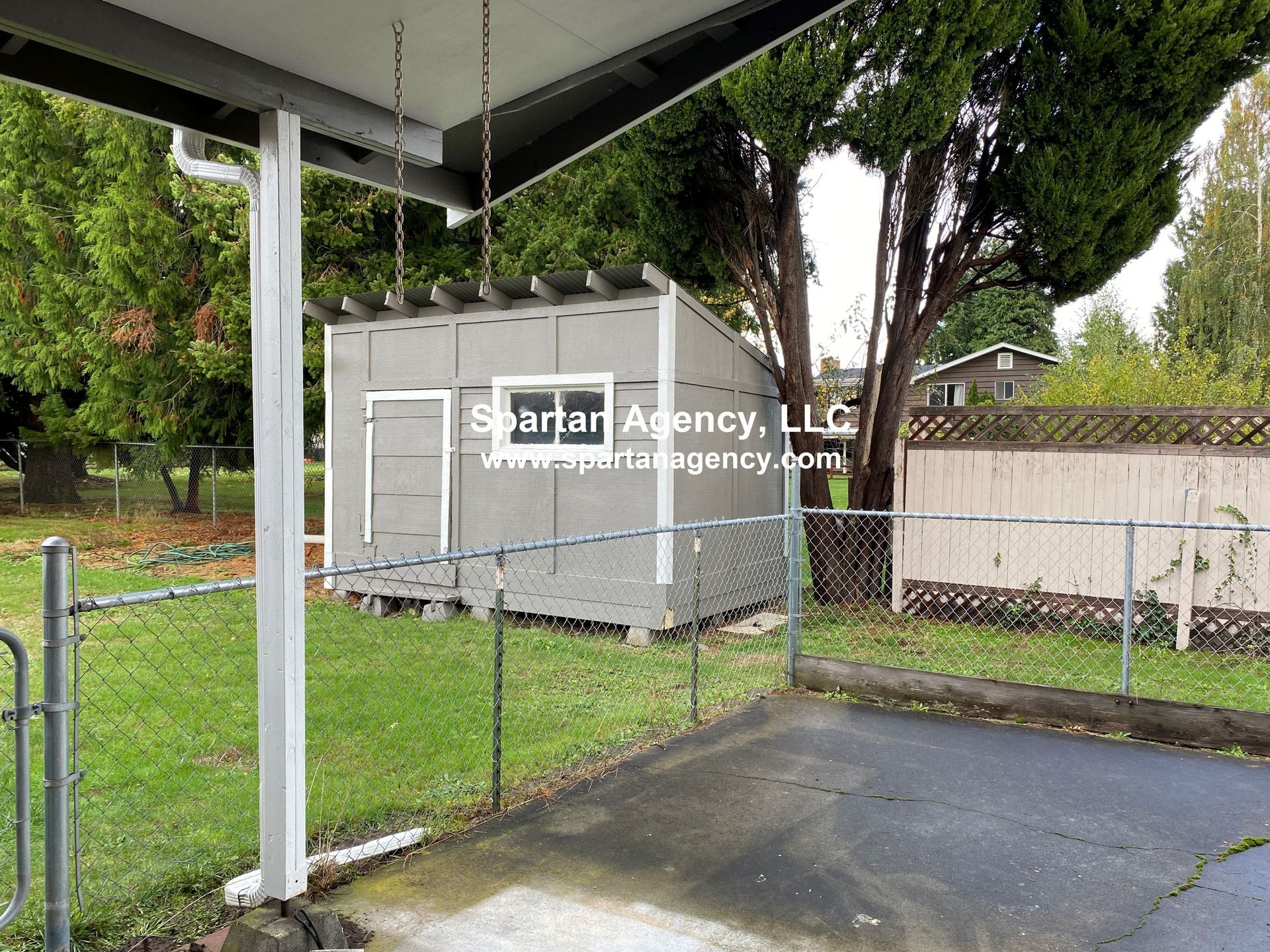 Backyard with a shed, a chain-link fence, and a concrete patio under a covered porch. Green grass and trees are visible.