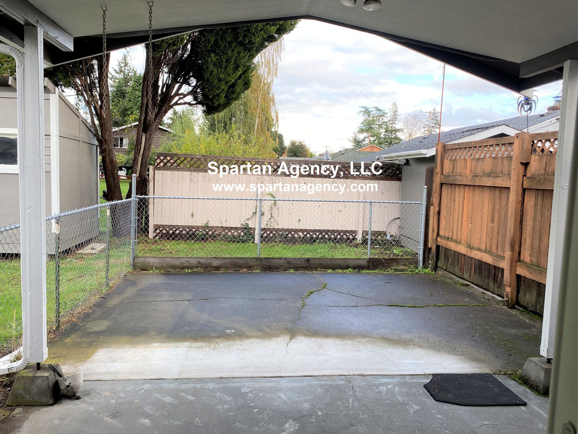 A concrete patio with a chain-link fence, wooden fence, and small building in the background. Cloudy sky visible.