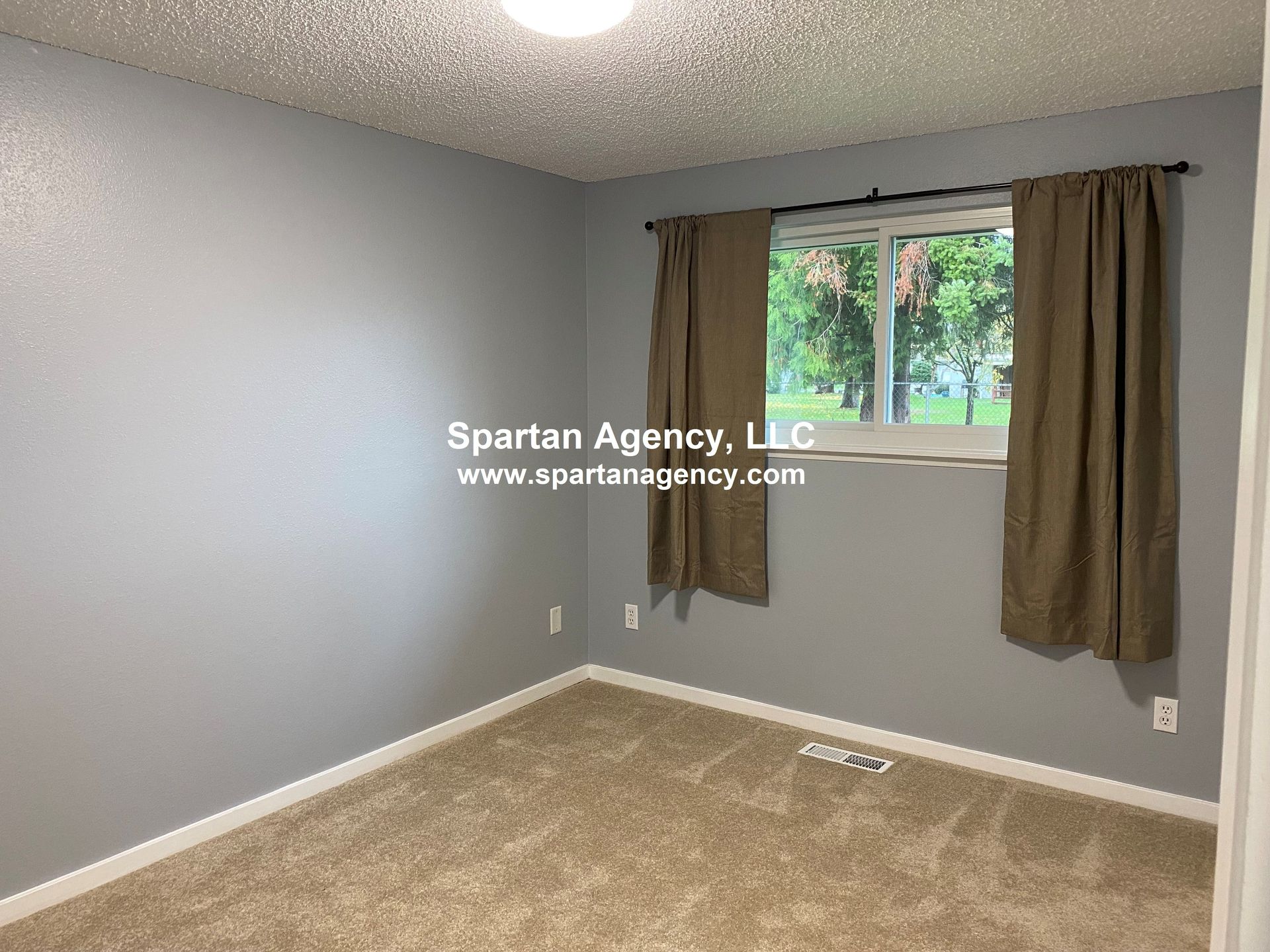 A small bedroom with gray walls, beige carpet, a window with brown curtains, and a white trim.