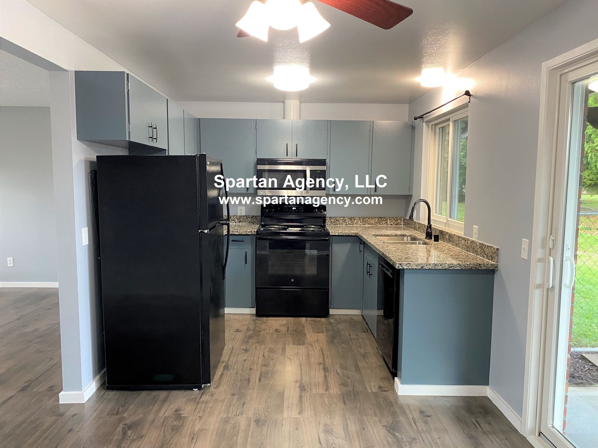 A kitchen with blue cabinets, black appliances, and granite countertops. A window to the right offers a view of a grassy yard.