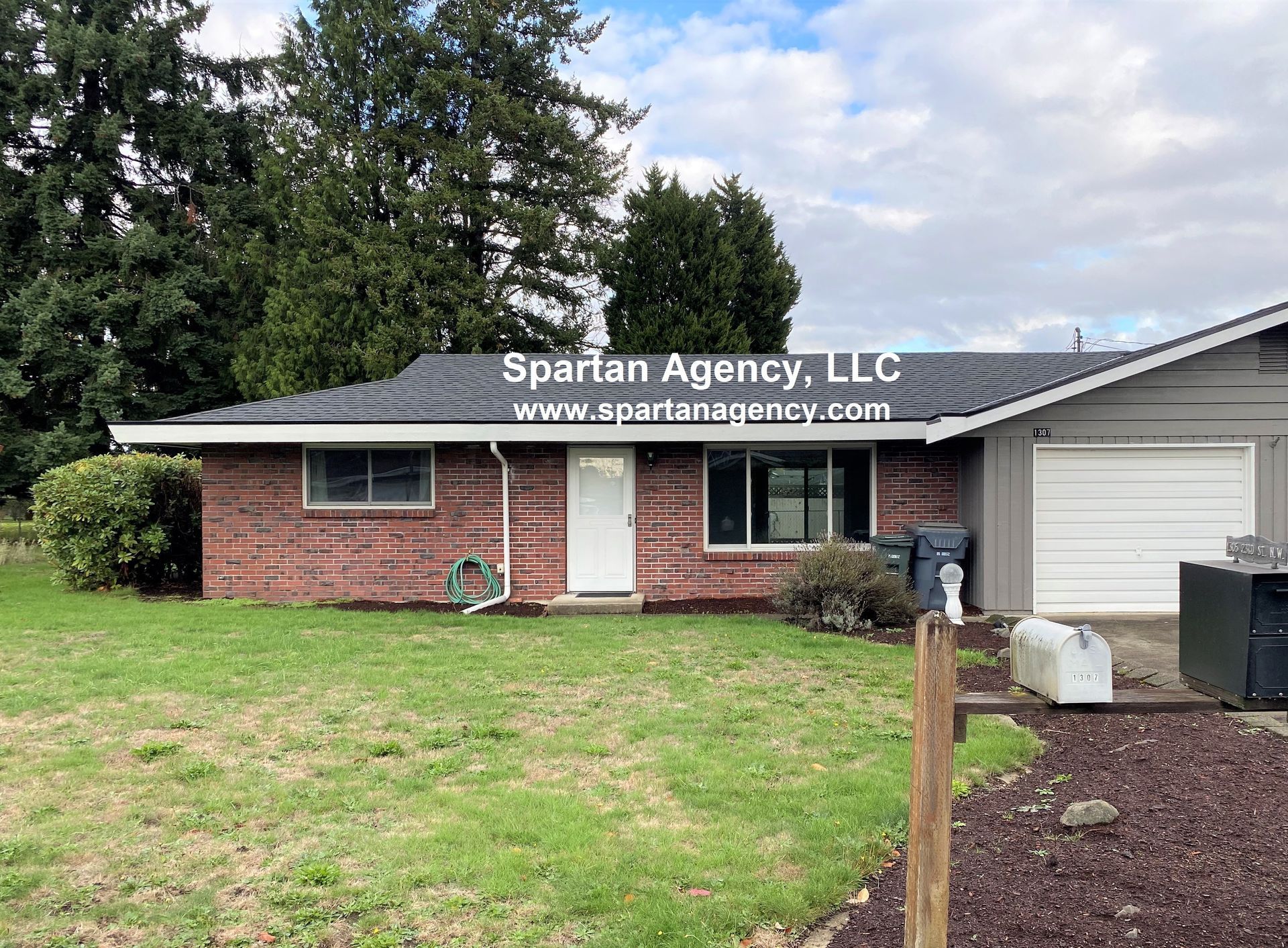 A brick ranch-style house with a gray garage door and white front door, on a green lawn. The Spartan Agency LLC logo is across the roof.