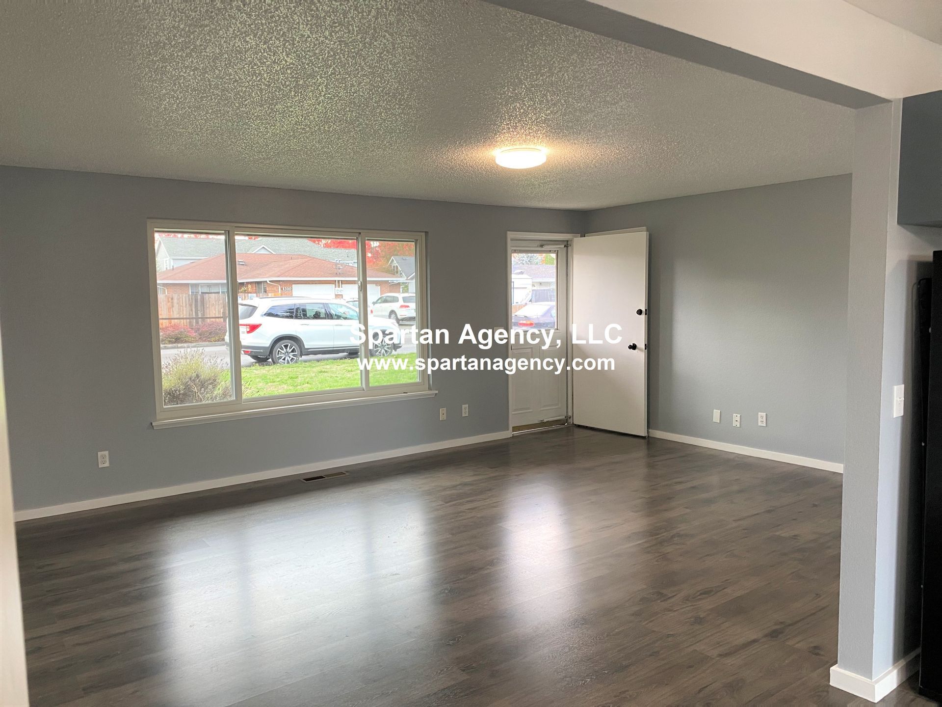Empty living room with gray walls, dark wood floors, and a large window overlooking a street. A white door is visible on the right.