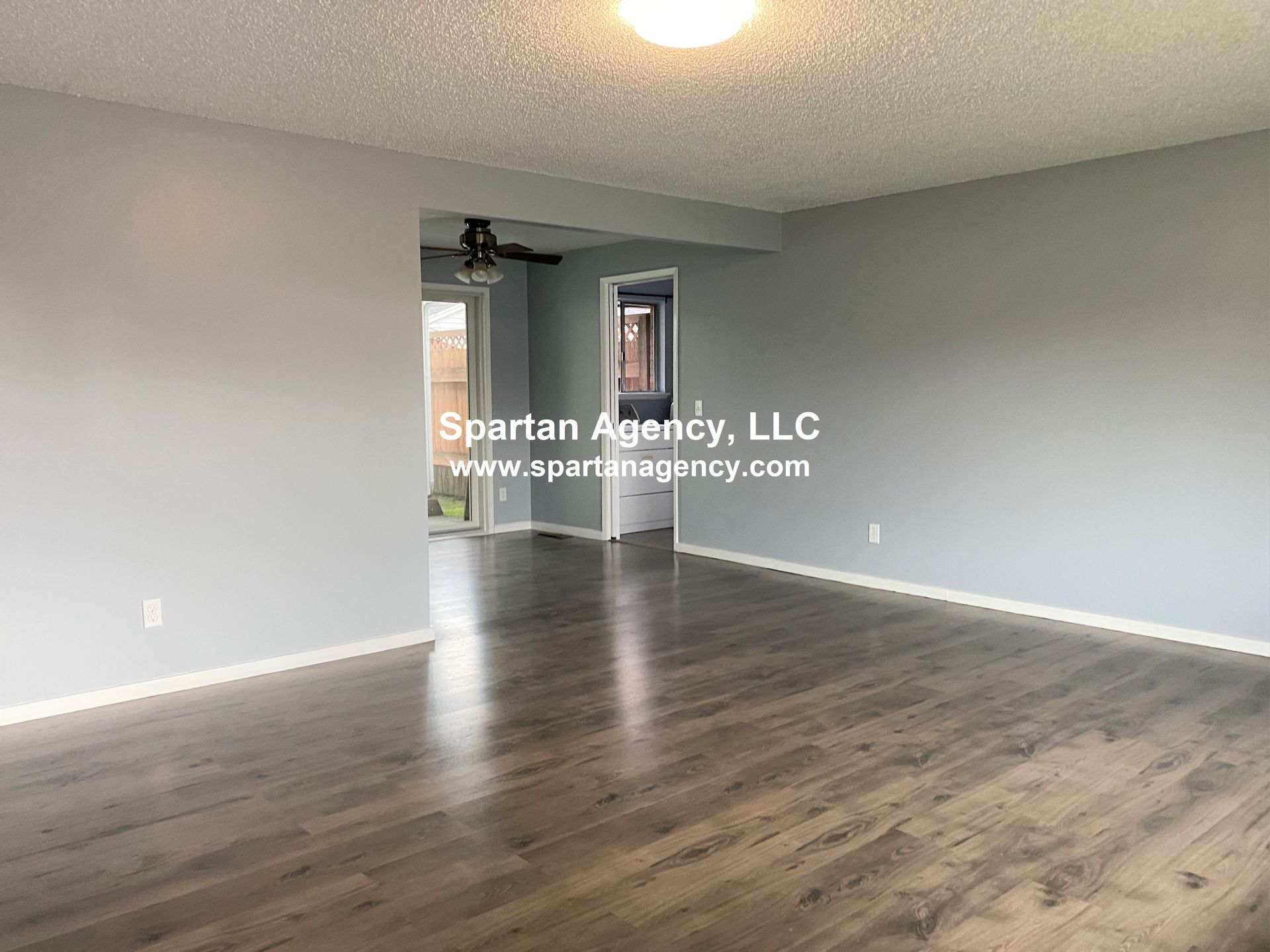 Empty living room with gray walls, wooden floors, and an entryway to another room. Spartan Agency watermark is visible.