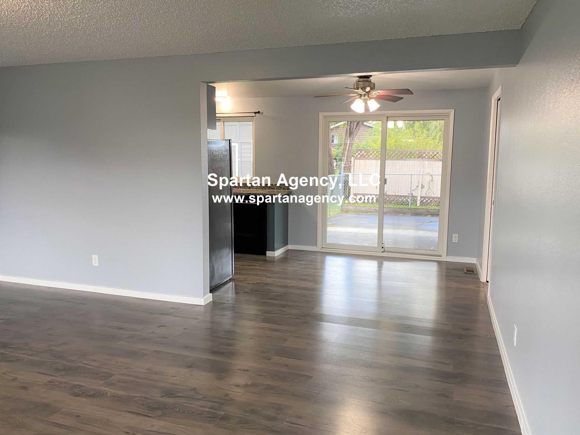 A room with gray walls, dark wood-look floors, and a sliding glass door. A kitchen is visible in the background.