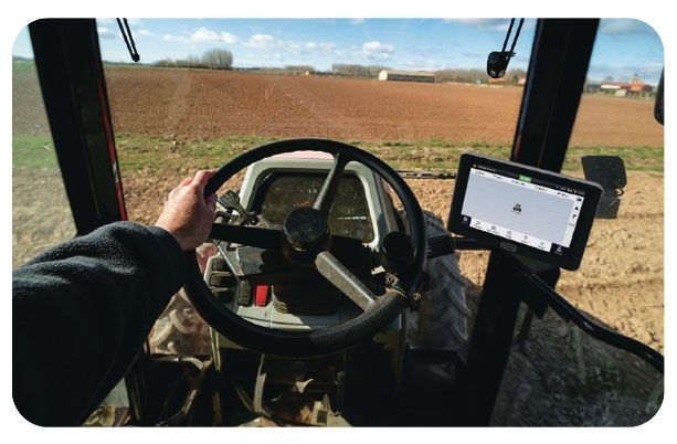 Hand on tractor steering wheel, view from inside the cab. GPS screen and farmland visible through the windshield.