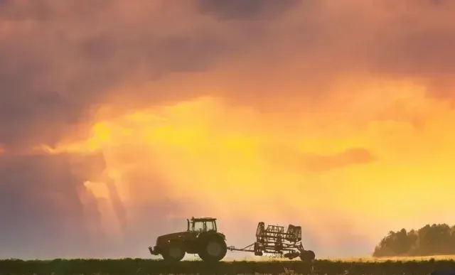 Tractor plowing a field at sunset; golden sky with clouds and trees in the distance.