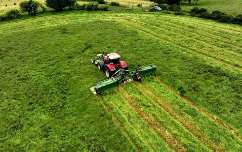 Red tractor mowing a large green field, creating parallel rows of cut grass.