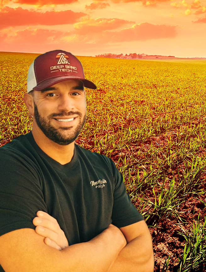 Man in cap and black shirt, arms crossed, smiling in a field with a sunset background.