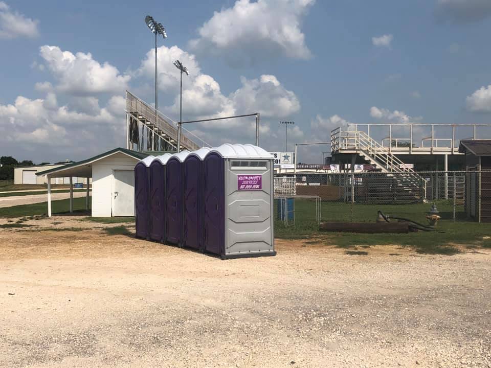 A row of purple portable toilets in front of bleachers and a white building.