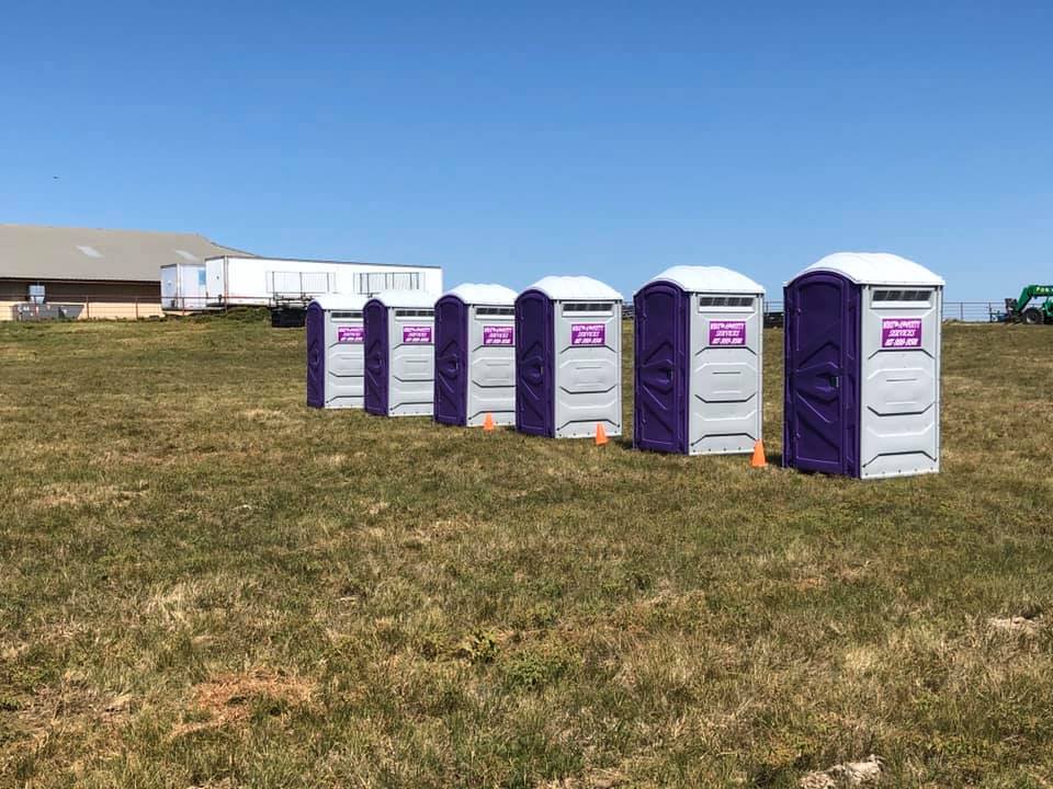 Six purple and gray portable toilets in a grassy field under a blue sky.