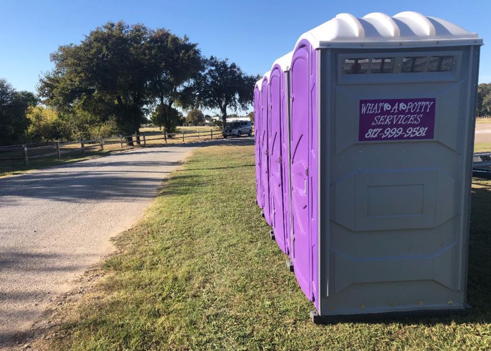 A row of purple portable toilets on a grassy area next to a paved path with trees in the background.