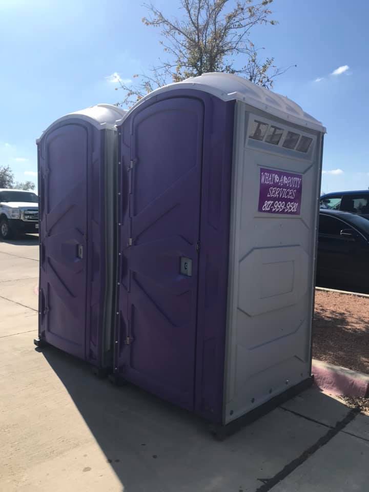 Two purple and gray portable toilets on a sidewalk in front of vehicles.