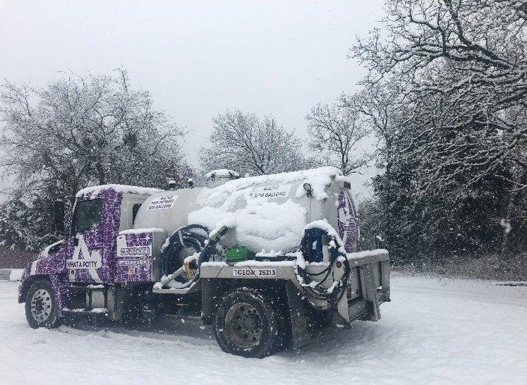 A snow-covered purple and white septic truck parked on a snowy road with leafless trees in the background.