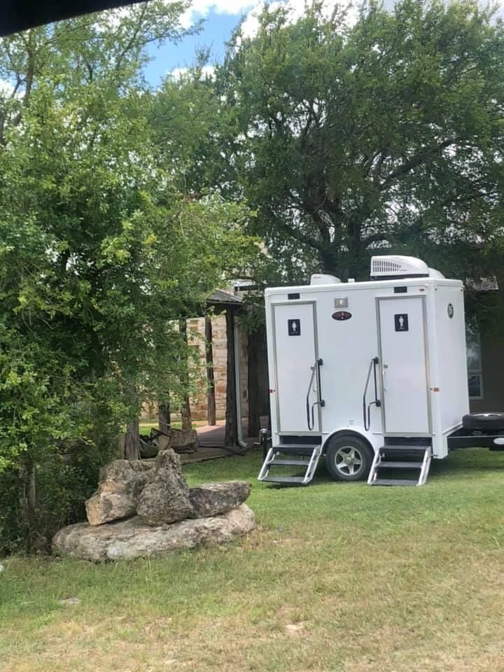 White portable restroom trailer on grass, two doors, steps, with trees and a rock.