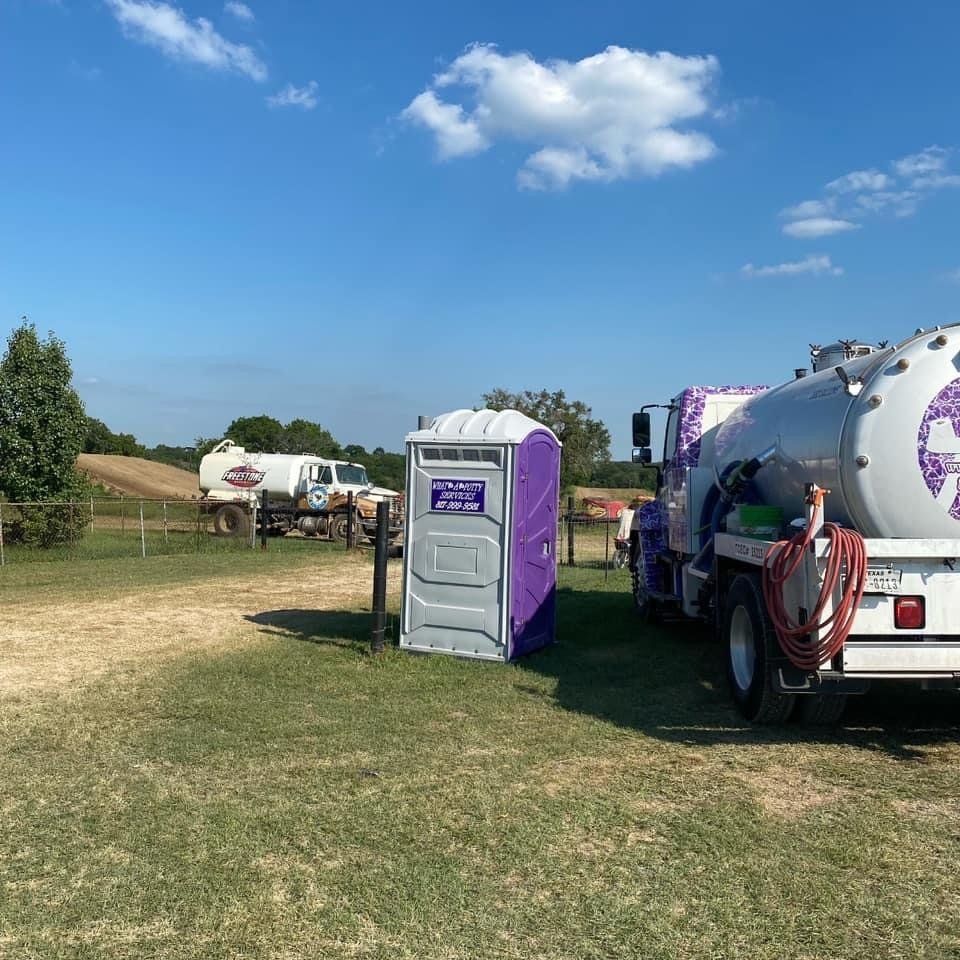 Portable toilet beside a truck, with another truck in the background, set in a grassy field under a blue sky.