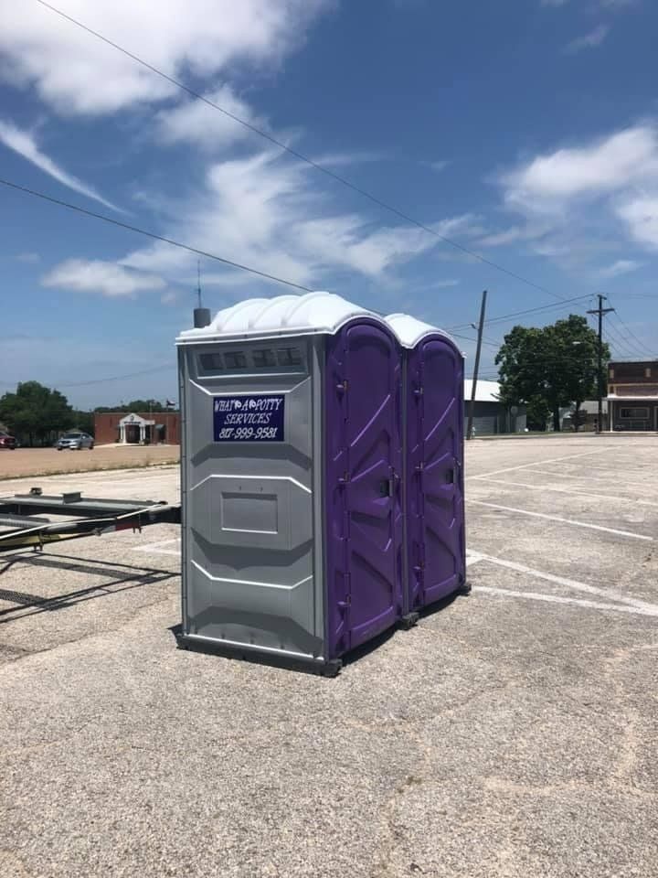 Two portable toilets, one gray and one purple, on a concrete lot under a blue sky with clouds.